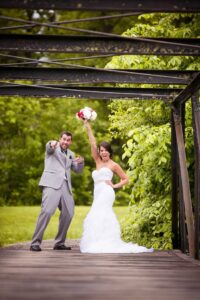 Bride in white dress raising bouquet, groom in gray suit pointing at the camera, both standing on a wooden bridge