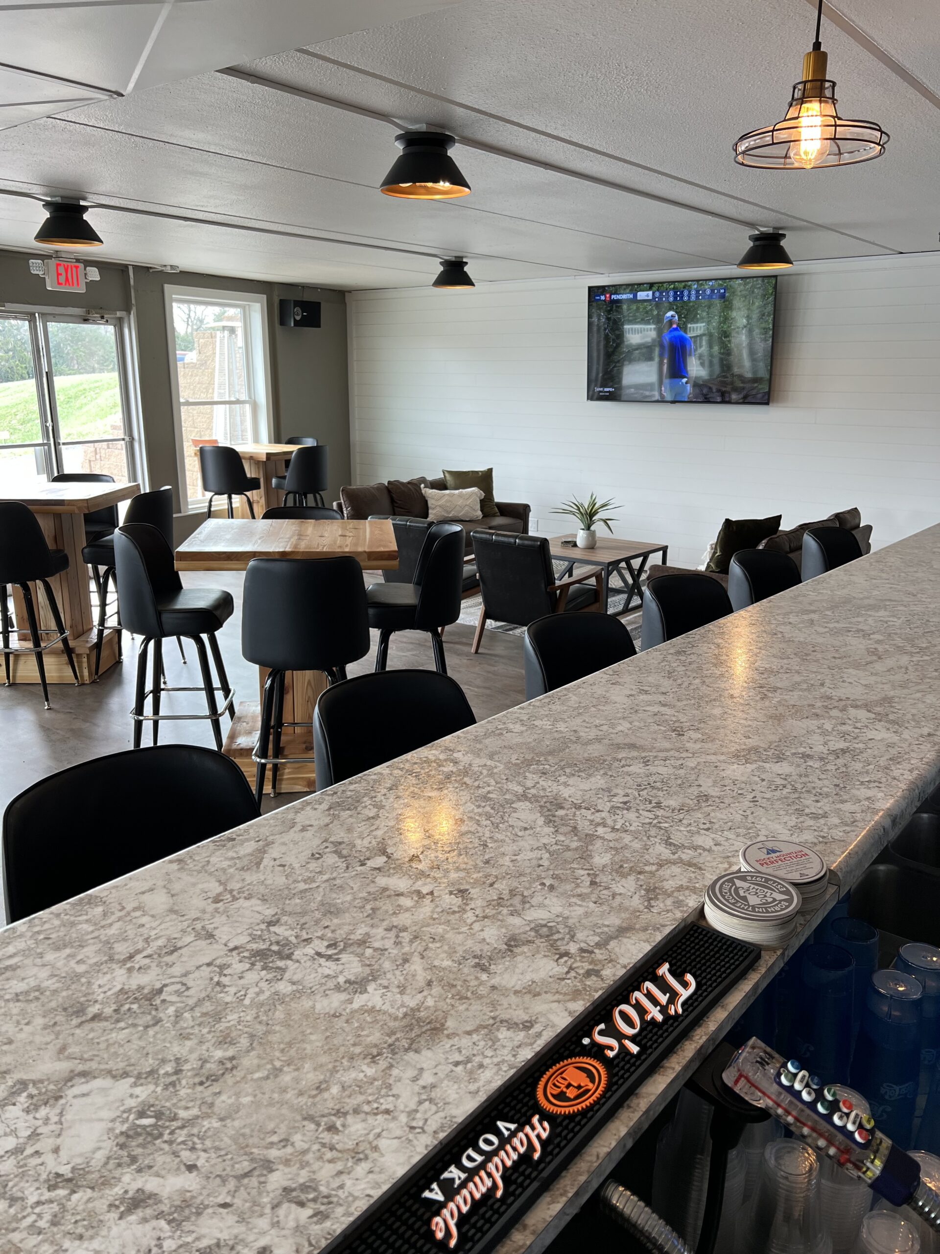 Modern bar interior with a granite counter in the foreground, high chairs, wooden tables, and a TV on the wall