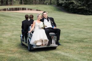 Bride and groom in wedding attire riding on the back of a golf cart