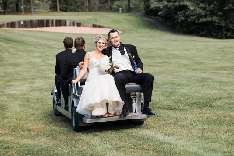 Bride and groom in wedding attire riding on the back of a golf cart