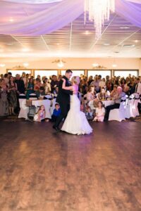 Bride and groom share their first dance on a wooden floor at their wedding, surrounded by seated and standing guests