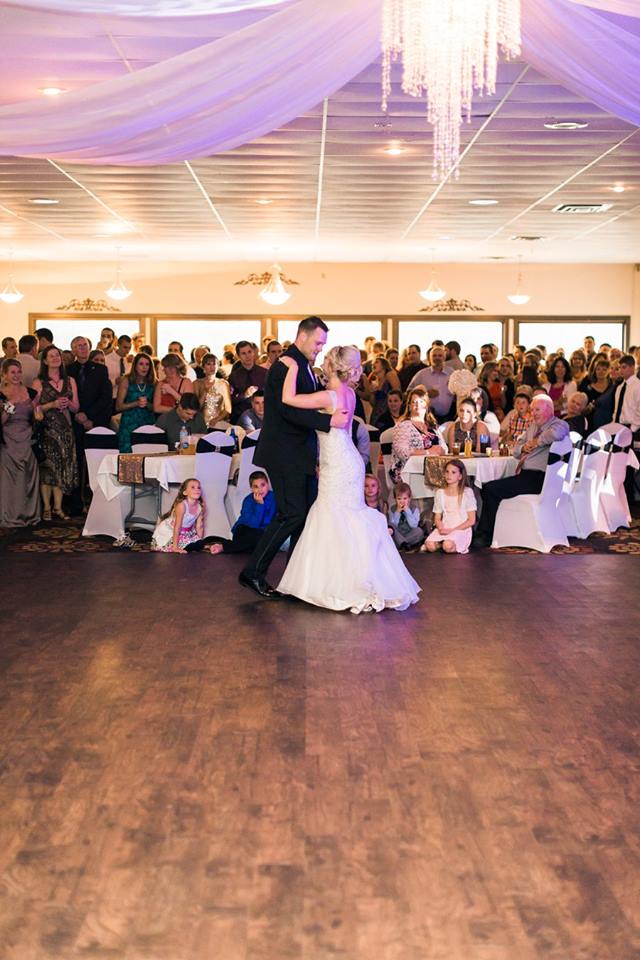Bride and groom share their first dance on a wooden floor at their wedding, surrounded by seated and standing guests