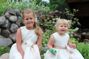 Two young girls in white dresses in front of a stone wall and flowers