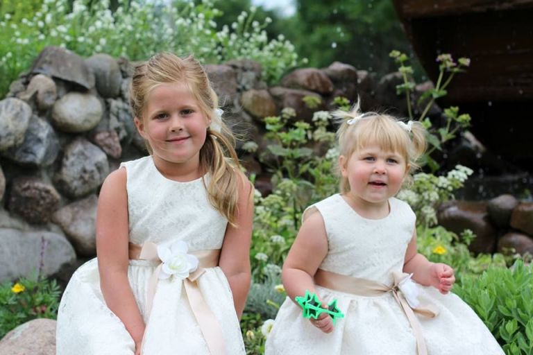 Two young girls in white dresses in front of a stone wall and flowers