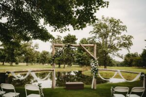Outdoor wedding setup by a pond with a wooden arch adorned in flowers