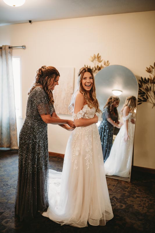 Bride in a white dress smiles joyfully as another woman helps adjust her dress
