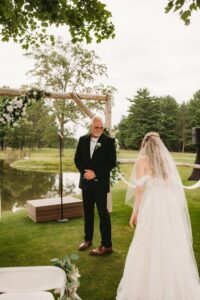 Bride in a white dress and veil approaching an older man in a suit under a floral arch