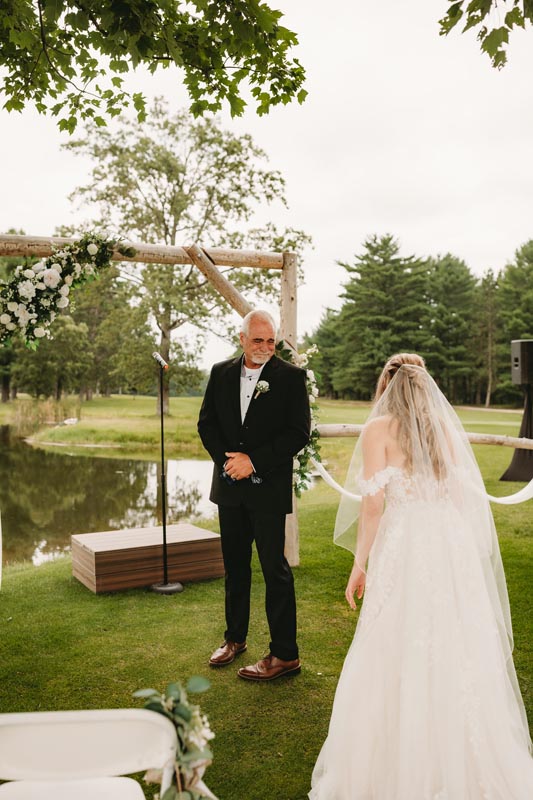 Bride in a white dress and veil approaching an older man in a suit under a floral arch