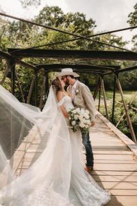 Bride in a white dress and groom in a cowboy hat kiss on a wooden bridge
