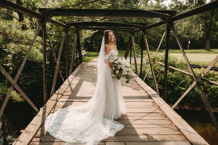 Bride in a flowing white dress and veil stands smiling on a wooden bridge, holding a bouquet of flowers