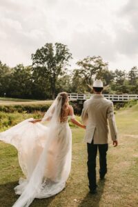 Bridge in a flowing dress and veil walking with the groom in a cowboy hat across a grassy field