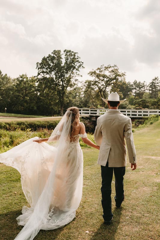 Bridge in a flowing dress and veil walking with the groom in a cowboy hat across a grassy field