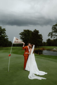 Newlywed couple sharing a kiss on a golf course with the groom holding a flag reading "The Andersons"