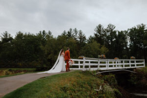 Bride in a white dress and groom in an orange suit crossing a bridge