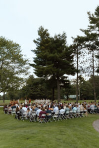 Outdoor wedding ceremony with guests seated in rows of chairs, surrounded by tall trees