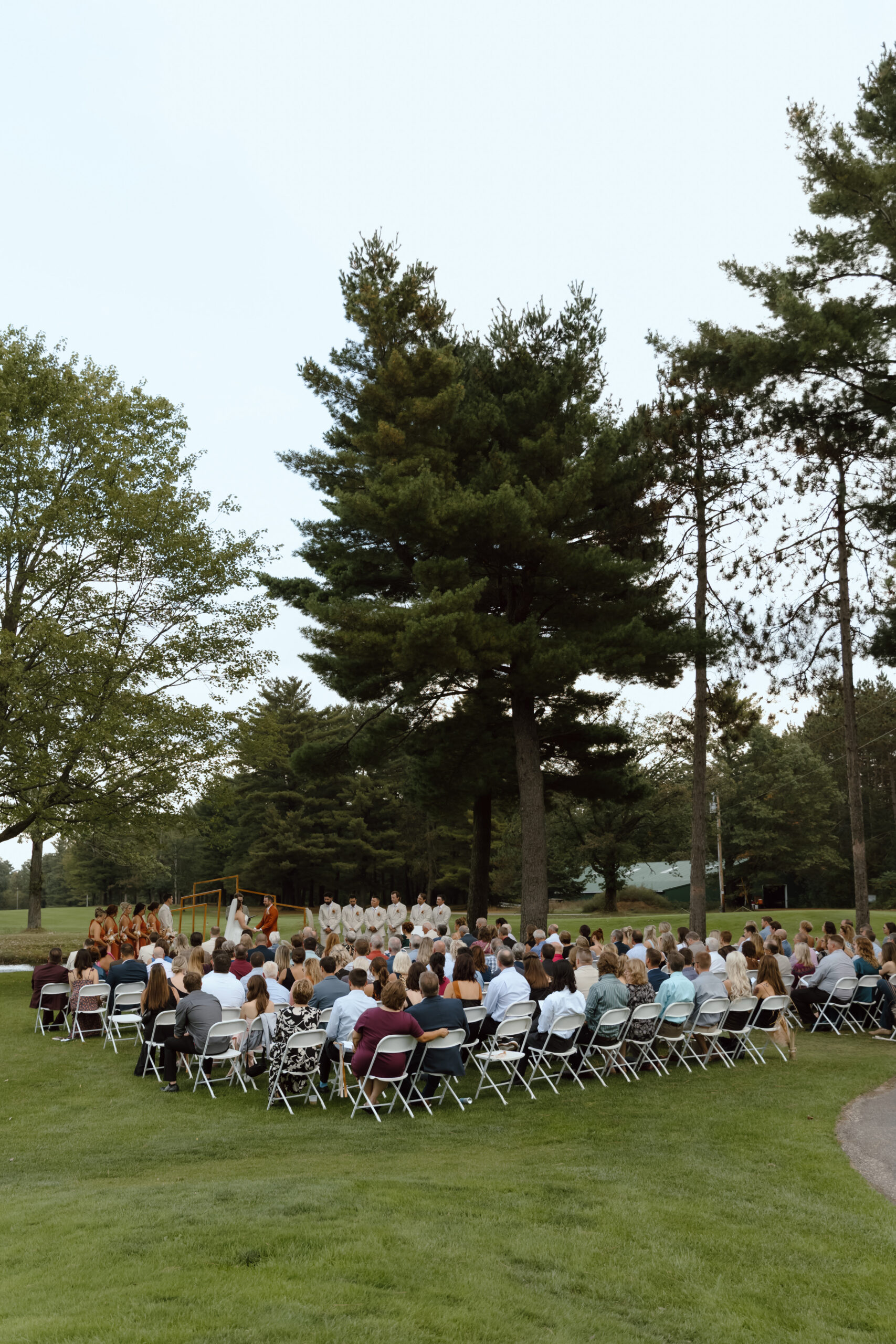 Outdoor wedding ceremony with guests seated in rows of chairs, surrounded by tall trees